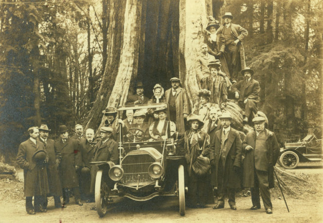 A large group of people pose with car in front of the Hollow Tree in Stanley Park. Photo taken in 1909.