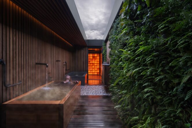 A woman relaxes in a cedar soaking tub next to ferns at Circle Wellness in Vancouver. There is no roof so she is looking up at the sky. 